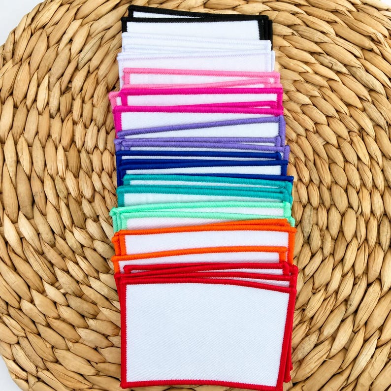 K&ouml;nnte beinhalten: Ein Stapel rechteckiger Stoffaufn&auml;her mit wei&szlig;er Mitte und gestickten R&auml;ndern in verschiedenen Farben wie Schwarz, Wei&szlig;, Pink, Lila, Blau, T&uuml;rkis, Gr&uuml;n, Orange und Rot, die in einer vertikalen S&auml;ule angeordnet sind.