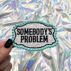 May include: Embroidered patch with the text "SOMEBODY'S PROBLEM" in black, set against a white background with a decorative floral design. The patch has a light blue border and a black outline. The patch is held up against a holographic background.