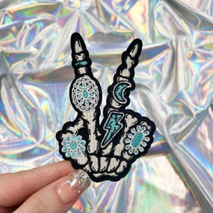 May include: A blue and white embroidered patch of a skeleton hand making the peace sign. The hand has a crescent moon, a lightning bolt, and floral designs on the fingers.