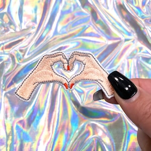 Pode incluir: Etiqueta bordada branca de duas mãos formando um coração com esmalte de unha vermelho.