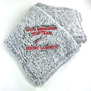 May include: A folded, gray, plush blanket with red embroidered text that reads "SOUTH WINNESHIEK TRAP TEAM" and "KENDRIC LANGRECK". A red rifle graphic is embroidered below the text. The blanket has a soft, fuzzy texture.