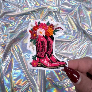 May include: Embroidered patch featuring a pair of pink cowboy boots with floral accents. The boots are adorned with white star details and topped with a bouquet of colorful flowers. The patch has a white border.