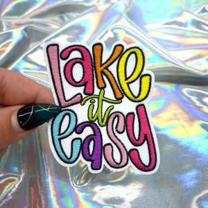 May include: Embroidered patch with the phrase "Lake it Easy" in colourful, bubble-style lettering. The patch features a white background with a black border, and the text is in shades of pink, orange, yellow, green, purple, and blue.