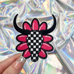 May include: A black and white checkered bull skull with pink flower petals around it. The patch is embroidered and has a white outline.
