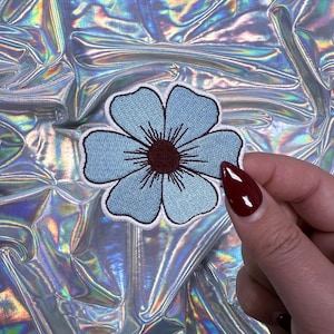 May include: A light blue embroidered flower patch with a white border. The flower has a dark maroon center and is set against a shiny, iridescent background. The patch is held by a person with red nail polish.