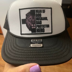 K&ouml;nnte beinhalten: Eine schwarz-wei&szlig;e Trucker-Cap mit einem rechteckigen Aufn&auml;her auf der Vorderseite. Der Aufn&auml;her zeigt ein schwarzes Kreuz, ein L&ouml;wengesicht und den Text: MAN OF GOD HUSBAND, DAD PROTECTOR HERO. Ein kleiner schwarzer Aufkleber auf dem Schirm zeigt OTTO WORLD FAMOUS FOAM FRONT OSFM TRUCKER.