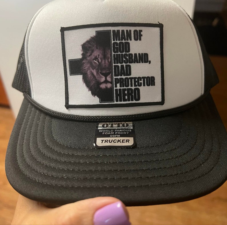 K&ouml;nnte beinhalten: Eine schwarz-wei&szlig;e Trucker-Cap mit einem rechteckigen Aufn&auml;her auf der Vorderseite. Der Aufn&auml;her zeigt ein schwarzes Kreuz, ein L&ouml;wengesicht und den Text: MAN OF GOD HUSBAND, DAD PROTECTOR HERO. Ein kleiner schwarzer Aufkleber auf dem Schirm zeigt OTTO WORLD FAMOUS FOAM FRONT OSFM TRUCKER.