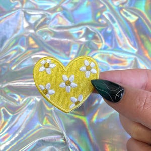 May include: A yellow heart-shaped patch embroidered with white and gold daisy-like flowers. The patch is held against a shimmering, iridescent background, creating a cheerful and decorative item.