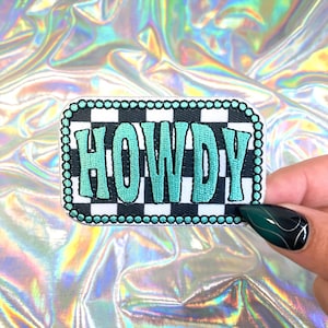 May include: A rectangular patch with the word "HOWDY" in turquoise, outlined in black, on a black and white checkered background. The patch is bordered with a string of small, silver-colored beads.
