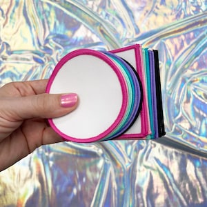 Puede incluir: Una pila de parches redondos, blancos y termoadhesivos, con costuras rosas, moradas, azules y verdes. Los parches están en blanco y listos para ser personalizados.