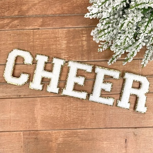 May include: White, textured letters spelling "CHEER" with gold trim, arranged diagonally on a wooden surface. A sprig of frosted greenery is in the upper right corner. The letters are likely for crafting or decoration.
