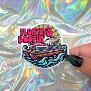 May include: Embroidered patch featuring a colorful pontoon boat design with the words "Floating & Boatin'" in pink and white. The patch has a white border and is set against a shimmering, iridescent background.