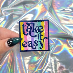 May include: A square embroidered patch with the words "take it easy" in navy blue script. The patch has a yellow border and a striped background in pastel colors. The background is a shiny, iridescent fabric.