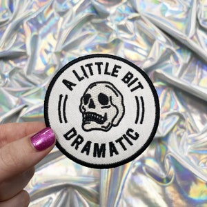 May include: White embroidered patch with a black skull and the text "A Little Bit Dramatic" in a black circle.