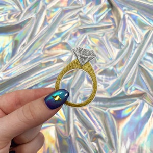 May include: Embroidered patch of a diamond engagement ring. The ring has a gold band with a white border and a silver diamond-shaped stone. The patch is designed to be sewn onto fabric.