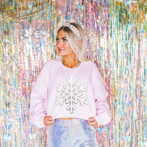 May include: A light pink cropped sweatshirt with a large white snowflake design. The model is wearing a sequined skirt and a jeweled headband. The background is a colorful, shimmering curtain.