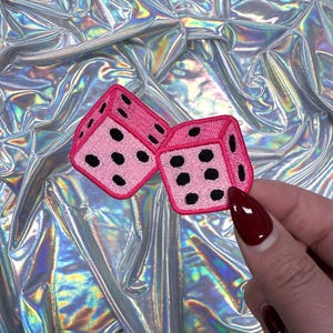 May include: Two pink dice patches with black dots against a shimmering, iridescent backdrop. The dice are embroidered with a black outline and feature a classic six-sided design. The patches are held by a hand with red nail varnish.