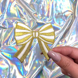 May include: A gold and white striped bow with a satin finish. The bow is embroidered with gold thread and has a white center.