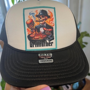 K&ouml;nnte beinhalten: Eine schwarz-wei&szlig;e Trucker-Cap mit einem rechteckigen Aufn&auml;her, der ein Skelett in Anzug und Fedora beim Grillen zeigt. Der Text Grillfather steht unter der Grafik. Ein schwarzer Aufkleber auf dem Schirm zeigt OTTO WORLD FAMOUS FOAM FRONT OSFM TRUCKER.