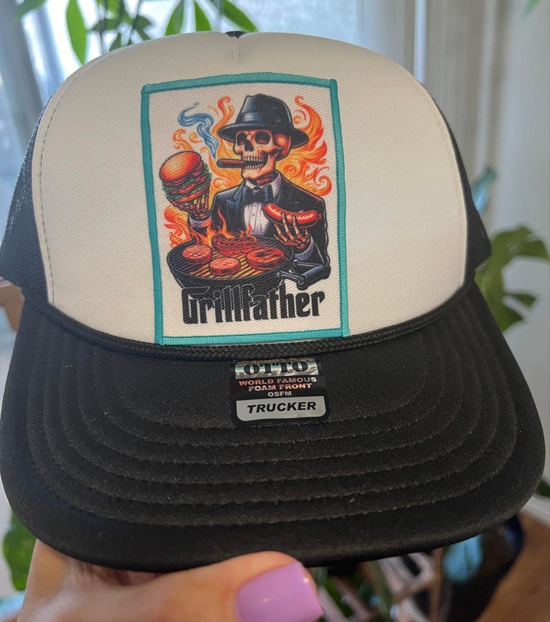 K&ouml;nnte beinhalten: Eine schwarz-wei&szlig;e Trucker-Cap mit einem rechteckigen Aufn&auml;her, der ein Skelett in Anzug und Fedora beim Grillen zeigt. Der Text Grillfather steht unter der Grafik. Ein schwarzer Aufkleber auf dem Schirm zeigt OTTO WORLD FAMOUS FOAM FRONT OSFM TRUCKER.