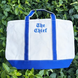 May include: A white canvas tote bag with blue accents. The bag features blue handles and a blue base. The words "The Chief" are embroidered in blue on the front of the bag. The bag is sitting on a bed of green leaves.