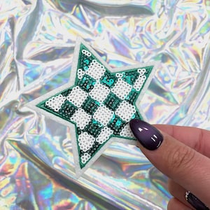 May include: A star-shaped patch with a green border and a checkered pattern of white and teal sequins. The patch is held against a shiny, iridescent background. The patch is a craft supply.