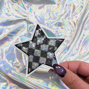 May include: A star-shaped patch with a black and silver checkered design, outlined in white. The patch is embellished with sequins and has a black border. The background is a shiny, iridescent material.