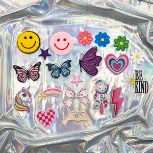May include: A collection of colorful embroidered patches featuring various designs. Includes a yellow smiley face, butterflies, flowers, hearts, a unicorn, a rainbow, a bow, a dreamcatcher, a lightning bolt, and the words "BE KIND".