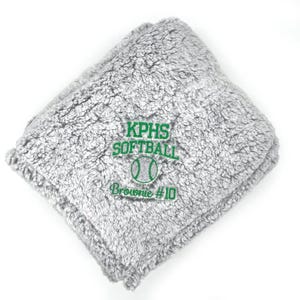 May include: A folded, gray, plush blanket with green embroidered text that reads "KPHS SOFTBALL" above a softball graphic and "Brownie #10". The blanket is made of a soft, fuzzy material.