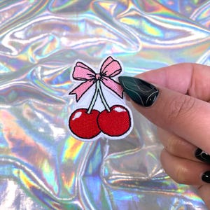 May include: Embroidered patch featuring two red cherries with white stems and a pink bow. The patch has a white border and is designed for application on clothing or accessories. The cherries are a vibrant red color.