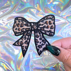 May include: A leopard print bow-shaped patch with a black outline. The patch features a tan background with brown spots, resembling a cheetah's pattern. The bow is held against a holographic background.
