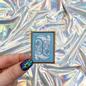 Könnte beinhalten: Ein blauer und goldener gestickter Aufnäher mit einem weißen Design aus drei stilisierten Stiefeln.