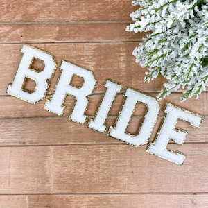 Puede incluir: Letras de fieltro blanco que deletrean "BRIDE" con ribete de purpurina dorada, dispuestas sobre una superficie de madera. Una ramita de vegetación cubierta de blanco se encuentra en la esquina superior derecha. Las letras son de estilo audaz.