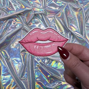 May include: Embroidered patch featuring a pair of pink lips with a white outline. The patch is held against a shiny, iridescent background. The lips are a vibrant pink color, with detailed stitching.