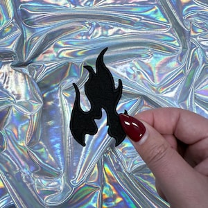 Pode incluir: Um patch de chama bordada preta. O patch é mantido contra um fundo brilhante e iridescente. O design da chama tem um contorno detalhado e é feito de um tecido texturizado.