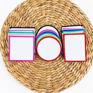 Può includere: Una collezione di sottobicchieri bianchi vuoti con bordi in tessuto colorato, disposti in tre pile. Due pile sono di forma quadrata con bordi vivaci in rosa, blu e verde, mentre la pila centrale è circolare con bordi rossi, arancioni e blu.