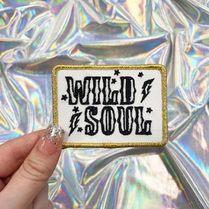 May include: A rectangular patch with the words "WILD SOUL" in black, stylized lettering. The patch has a white background with a gold border and small star and lightning bolt accents. The patch is held up against a shiny, iridescent background.