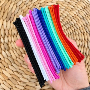 K&ouml;nnte beinhalten: Eine Sammlung d&uuml;nner, elastischer Haargummis, angeordnet in einem Farbspektrum aus Schwarz, Wei&szlig;, Pink, Lila, Dunkelblau, T&uuml;rkis, Orange und Rot. Diese Haaraccessoires sind flexibel, gekordelt und weisen eine glatte, gleichm&auml;&szlig;ige Textur auf.