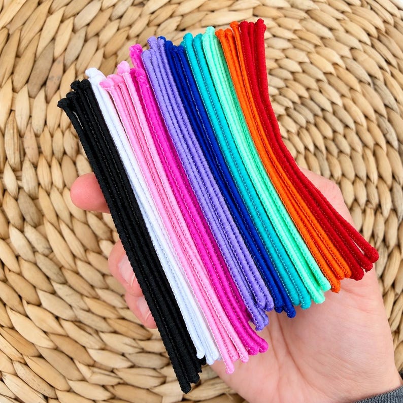 K&ouml;nnte beinhalten: Eine Sammlung d&uuml;nner, elastischer Haargummis, angeordnet in einem Farbspektrum aus Schwarz, Wei&szlig;, Pink, Lila, Dunkelblau, T&uuml;rkis, Orange und Rot. Diese Haaraccessoires sind flexibel, gekordelt und weisen eine glatte, gleichm&auml;&szlig;ige Textur auf.