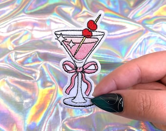 May include: Embroidered patch featuring a pink cocktail in a martini glass, adorned with a cherry garnish. The patch includes a pink bow detail and star accents. The design is outlined in black and white.