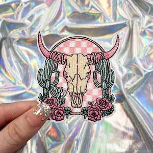 May include: Embroidered patch featuring a pink and white checkered background with a white cow skull, surrounded by green cacti and pink flowers.