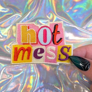 May include: Embroidered patch with the words "hot mess" in colorful, block letters. The patch features a pink and yellow background with orange, purple, and white lettering. The patch is on a holographic background.