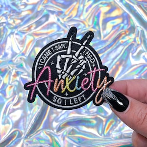 May include: Black and white embroidered patch featuring a skeleton hand making the peace sign. The text around the hand reads "I came I saw I had so I left" and the word "Anxiety" is written in rainbow colours.