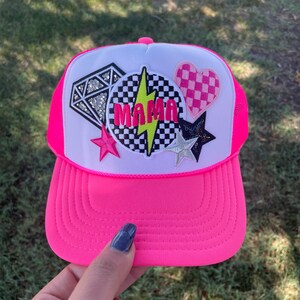 May include: A pink and white trucker hat with a checkered and lightning bolt patch that says "MAMA" in yellow letters. The hat also has a diamond, heart, and star patches.