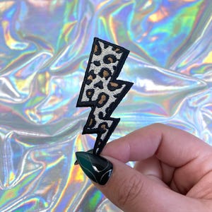 Pode incluir: Um patch em forma de raio com estampa de leopardo e bordas pretas. O patch apresenta um padrão de estampa animal em tons de bege e marrom. O patch está sobre um fundo brilhante e iridescente.