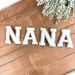 Könnte beinhalten: Weiße Filzbuchstaben, die "NANA" buchstabieren, mit goldfarbenem Glitzerbesatz. Die Buchstaben befinden sich auf einem Holzgrund.