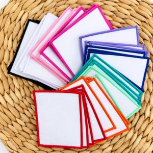 May include: A collection of rectangular fabric patches with white centers and stitched borders in various colors including black, pink, purple, blue, teal, green, orange, and red, arranged in a fanned-out display on a woven surface.