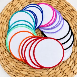 Könnte beinhalten: Eine Sammlung runder weißer Stoffaufnäher mit gestickten Rändern in verschiedenen Farben wie Rot, Orange, Hellgrün, Türkis, Blau, Lila, Pink und Schwarz, die in einem fächerartigen, überlappenden Muster auf einer geflochtenen Oberfläche angeordnet sind.