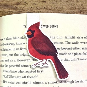May include: A red cardinal bookmark with the words "Saved Books" printed on it. The bookmark is placed on a page of an open book.