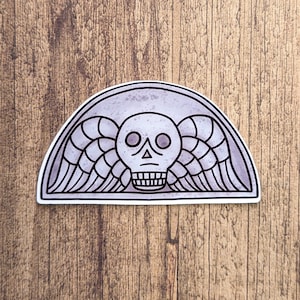 May include: A sticker with a gray background and a black and white illustration of a skull with wings. The skull has two eye sockets and a mouth with teeth. The wings are spread out on either side of the skull.